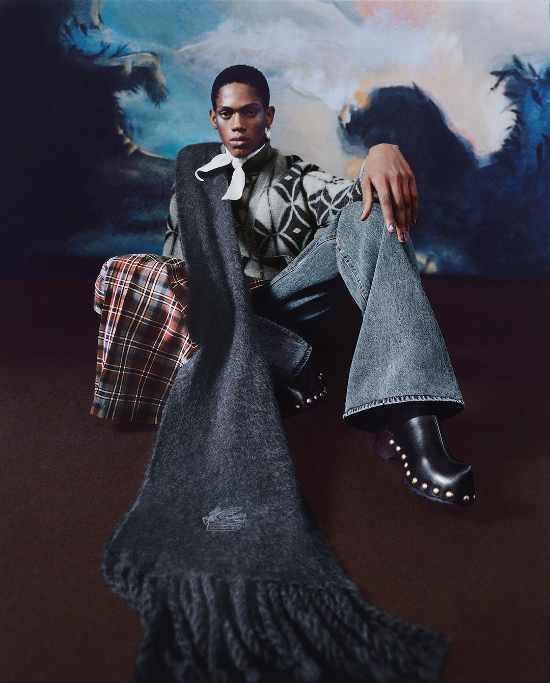 An African American male model sits in stylish denim and clogs, accessorized with a scarf, on a dark brown surface with a cloud backdrop.