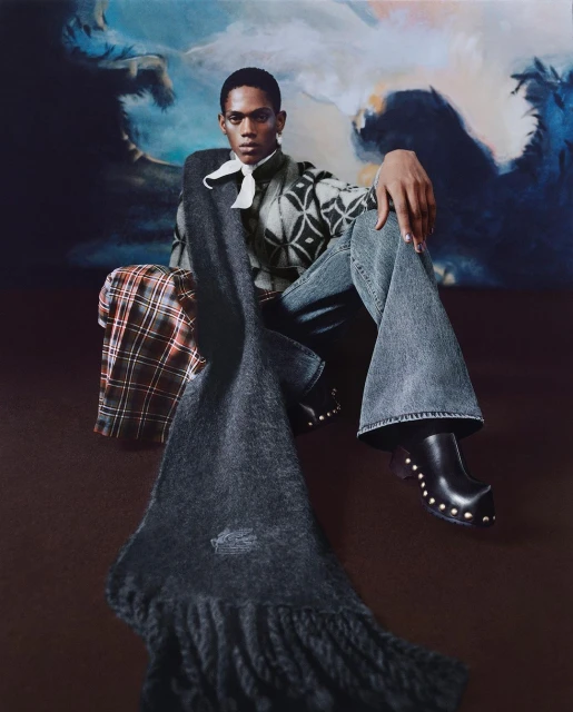 An African American male model sits in stylish denim and clogs, accessorized with a scarf, on a dark brown surface with a cloud backdrop.