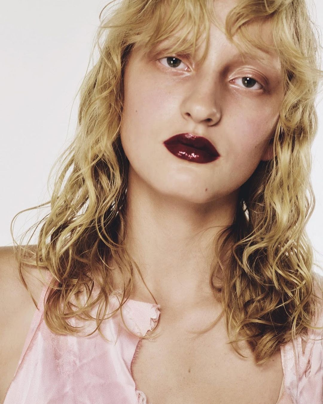 Close-up portrait of a woman with blonde wavy hair, glossy dark red lips, and a light pink top against a white seamless background.