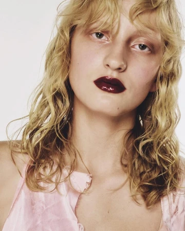 Close-up portrait of a woman with blonde wavy hair, glossy dark red lips, and a light pink top against a white seamless background.