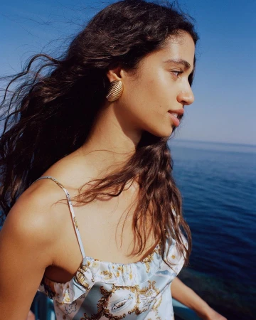 Profile of a woman with curly hair and gold earrings in a light blue dress, set against a blue sky and ocean backdrop.