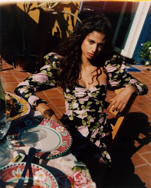 Woman in a floral dress sits at an outdoor table with plates and drinks, posing for a fashion shot on a brick patio with natural light.