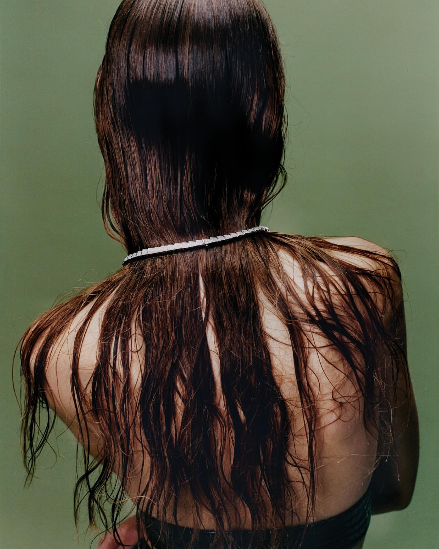 Back view of a woman with wet dark brown hair cascading down her back, accented by a delicate pearl necklace, against a pale green backdrop.