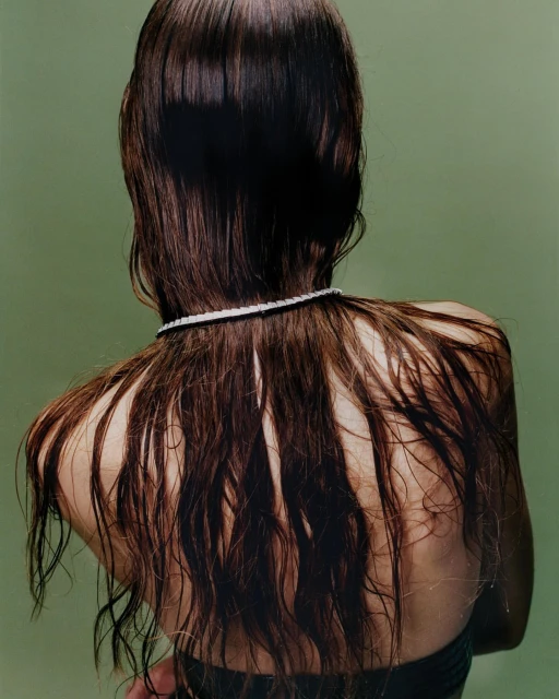 Back view of a woman with wet dark brown hair cascading down her back, accented by a delicate pearl necklace, against a pale green backdrop.