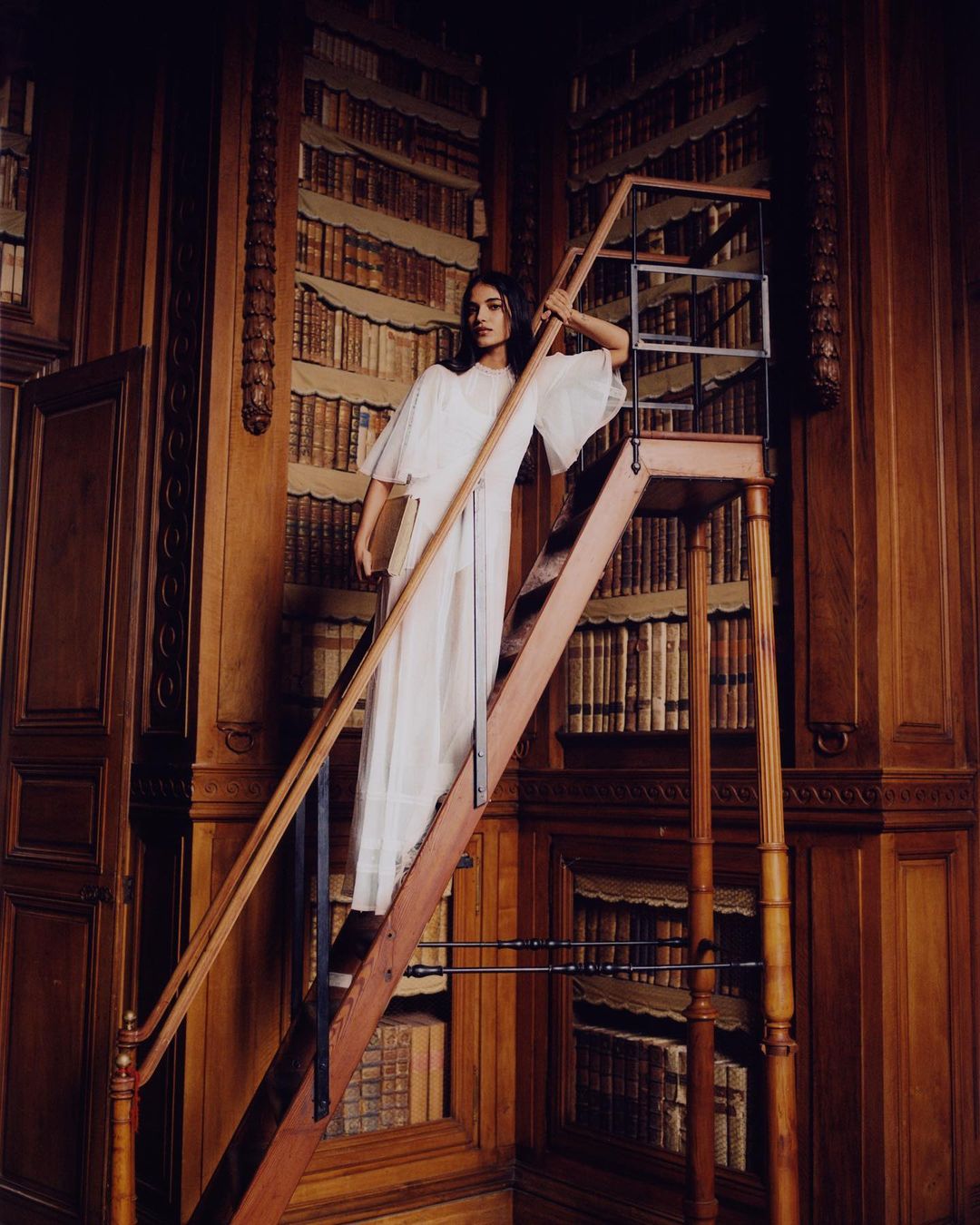 Woman in a white dress standing on a rolling ladder in a library with floor-to-ceiling bookshelves filled with antique books.
