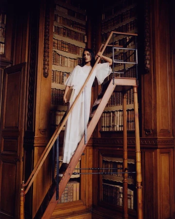 Woman in a white dress standing on a rolling ladder in a library with floor-to-ceiling bookshelves filled with antique books.