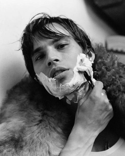 Young adult man with shaving cream on his face, holding a straight razor, with a fur covering around his shoulders in a black and white portrait.