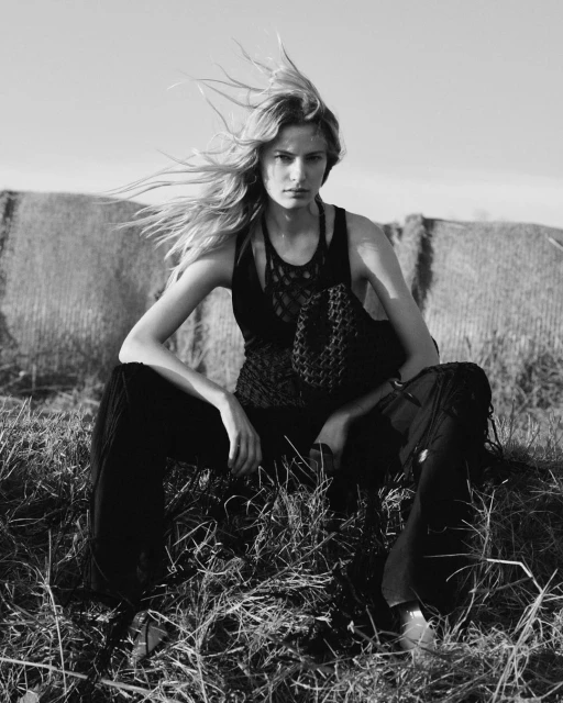 Black and white portrait of a female fashion model with long hair sitting in a grassy field with hay bales in the background.