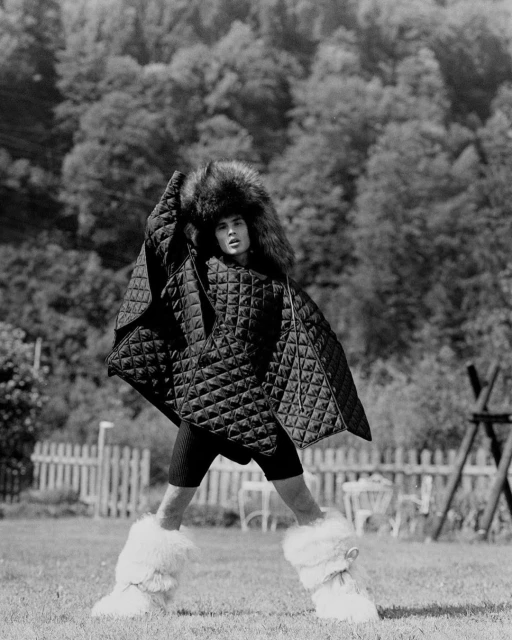Monochrome shot of a person in a furry hat, quilted poncho and furry boots posing on green grass in front of a white picket fence.