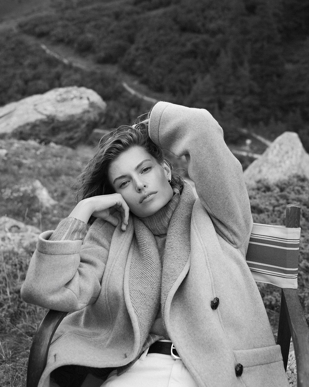 Woman in coat poses on a chair outdoors with mountains and foliage. Neutral tones, black and white. Fashion, travel, lifestyle.