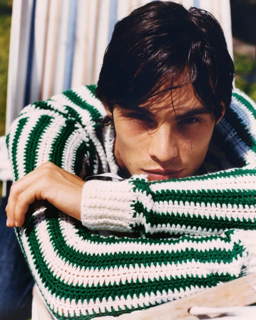 Close-up shot of a young man with dark hair, fair skin, and a striped knit sweater posing outdoors in a natural light setting.
