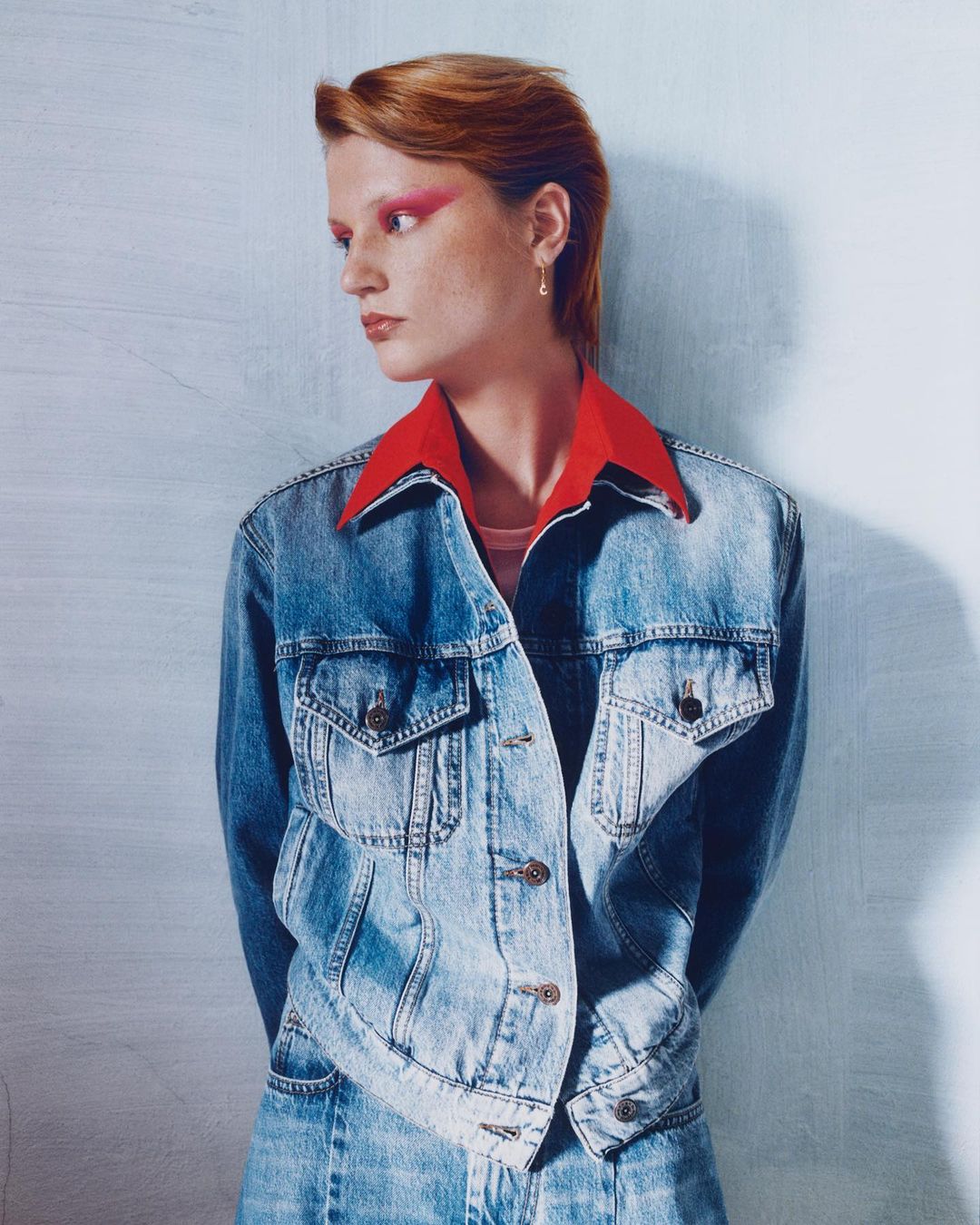 Portrait of a female model in a denim jacket and matching jeans, posing in front of a textured wall, with red eyeshadow.