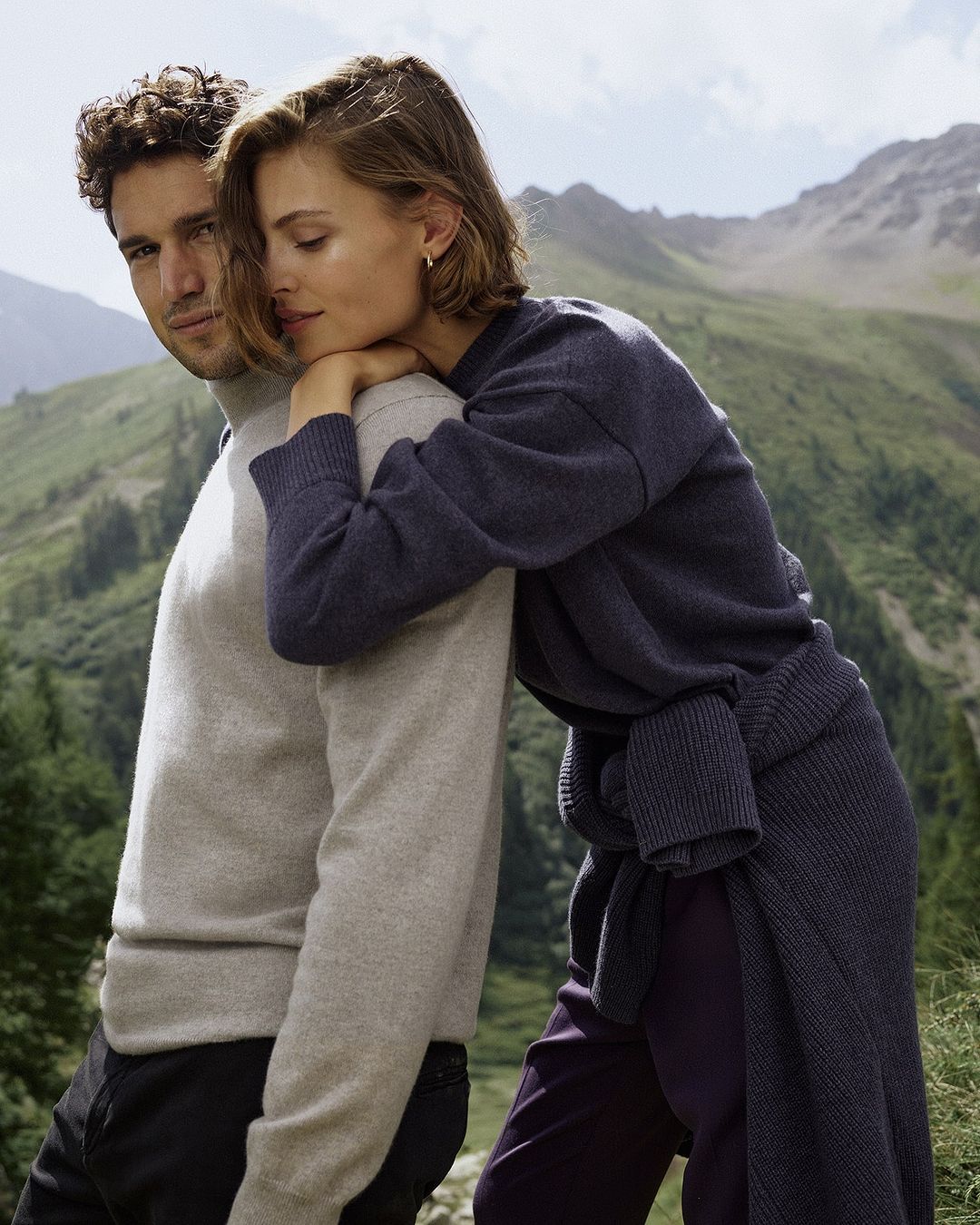 Couple posing outdoors with scenic mountain backdrop. The female leans on the male, both wearing knit sweaters, captured in soft, natural lighting.