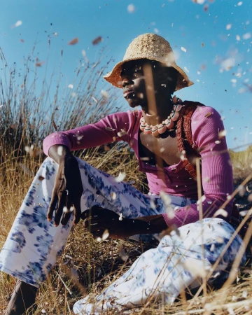 Man wearing a straw bucket hat, pink top, and blue patterned trousers sitting outdoors amongst tall grass under a clear blue sky