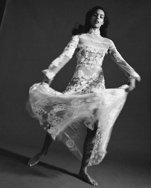 Elegant woman in sheer lace gown poses in a studio, capturing a blend of fashion and fine art with a monochromatic palette.