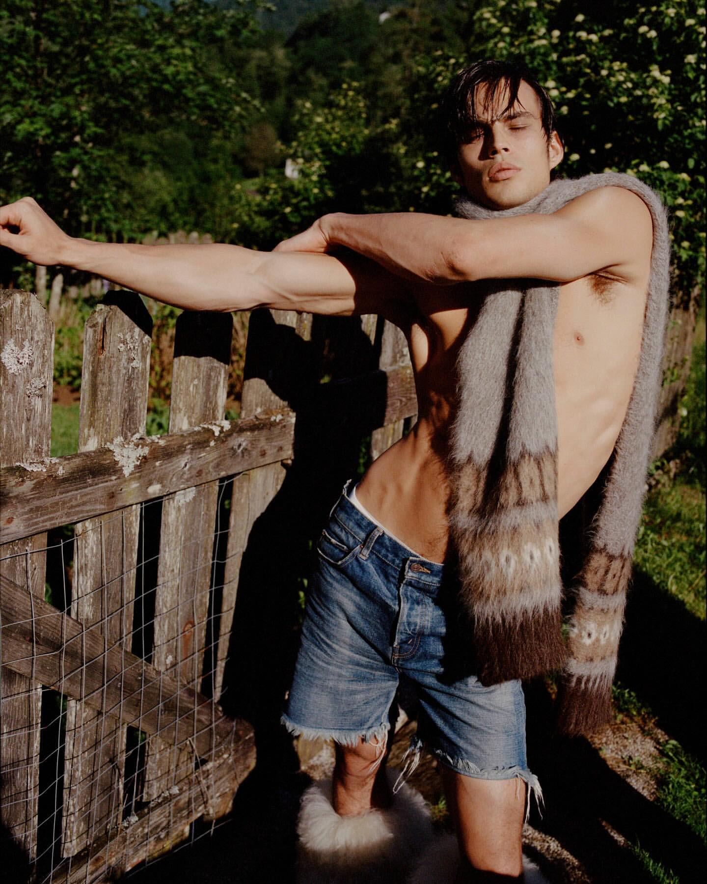 Attractive adult male with bare torso poses with a scarf draped around his neck in an outdoor setting with a wooden fence.