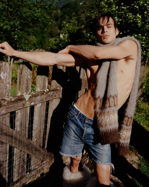Attractive adult male with bare torso poses with a scarf draped around his neck in an outdoor setting with a wooden fence.