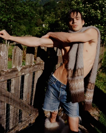 Attractive adult male with bare torso poses with a scarf draped around his neck in an outdoor setting with a wooden fence.