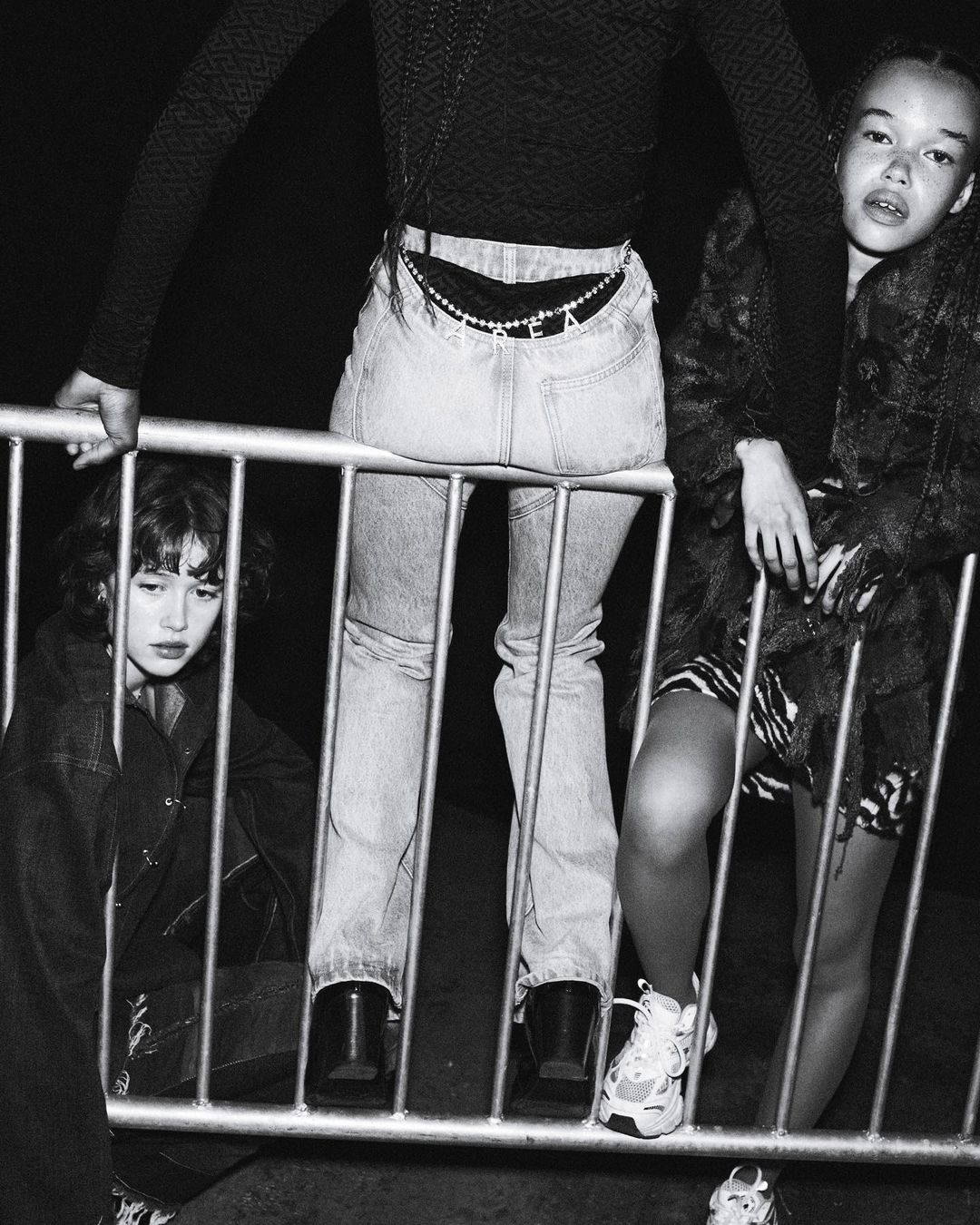 Three young women in a black and white fashion photo. One sits behind a metal barrier, one leans on it, and one stands next to it.