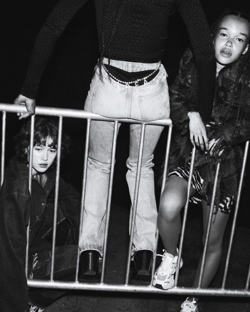 Three young women in a black and white fashion photo. One sits behind a metal barrier, one leans on it, and one stands next to it.