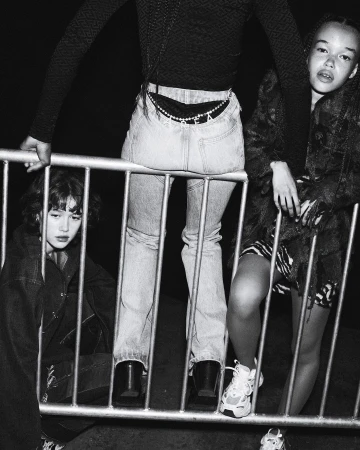Three young women in a black and white fashion photo. One sits behind a metal barrier, one leans on it, and one stands next to it.