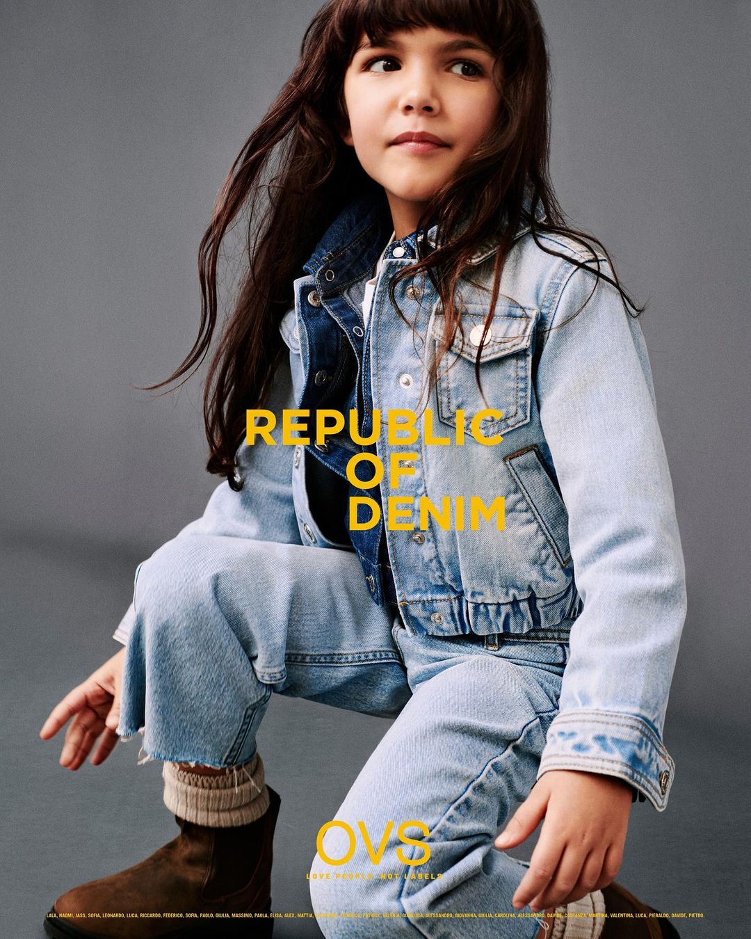 A young child models denim jacket and jeans with brown suede boots and striped socks against a grey background, fashion editorial.
