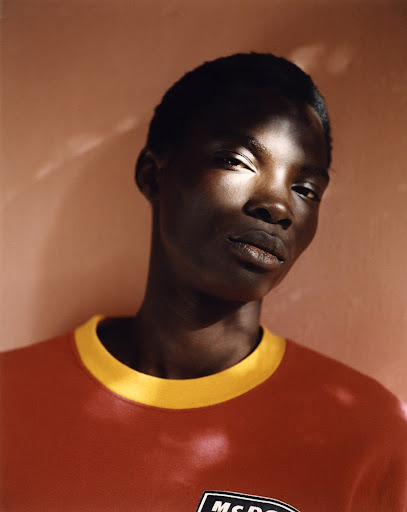 Portrait of a dark-skinned adult male with a close-cropped haircut wearing a red and yellow t-shirt against a reddish-tan wall.