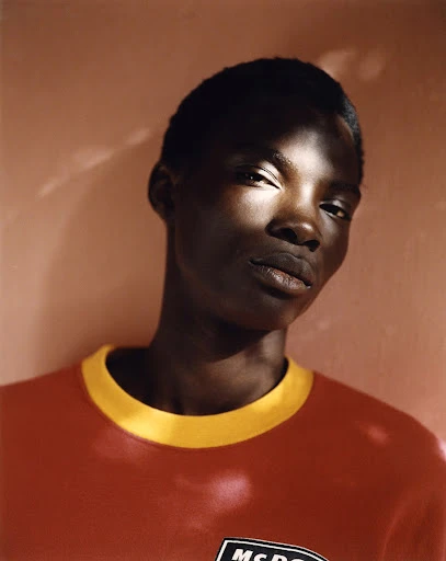 Portrait of a dark-skinned adult male with a close-cropped haircut wearing a red and yellow t-shirt against a reddish-tan wall.