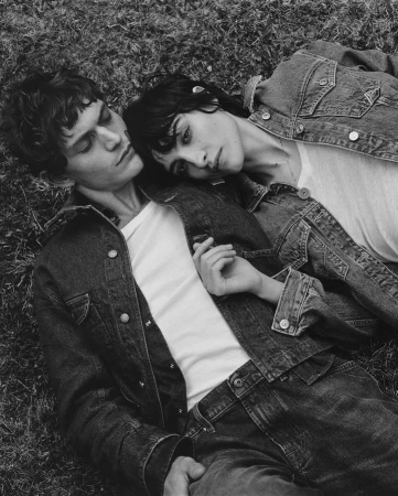 Monochrome portrait of a young man and woman in denim jackets lying together on grassy ground.