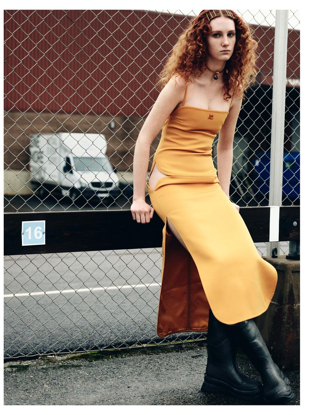 A redhead woman sits on a railing in mustard yellow dress and black boots, brick building and white van in the background.