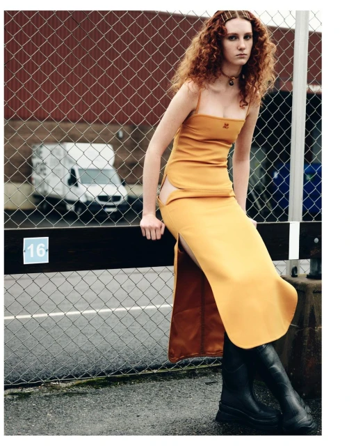 A redhead woman sits on a railing in mustard yellow dress and black boots, brick building and white van in the background.
