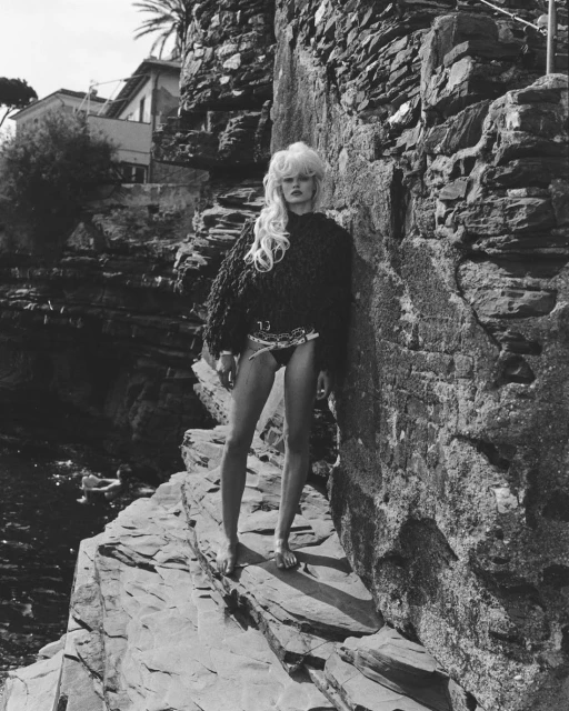 A woman with blonde hair stands barefoot on a rocky outcrop near the sea, wearing a dark sweater and swimsuit bottoms in a black-and-white fashion shot.