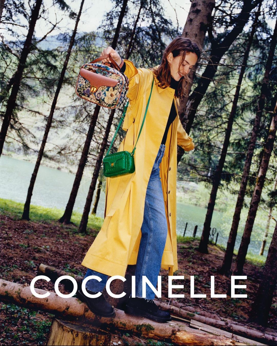 A stylish woman poses outdoors wearing a yellow coat and blue jeans, carrying two Coccinelle handbags in a forest setting.