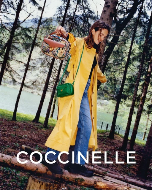 A stylish woman poses outdoors wearing a yellow coat and blue jeans, carrying two Coccinelle handbags in a forest setting.