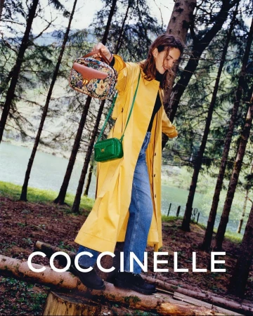 A stylish woman poses outdoors wearing a yellow coat and blue jeans, carrying two Coccinelle handbags in a forest setting.