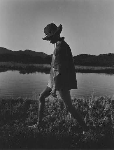 Model wearing a wide-brimmed hat and dark jacket walking by a calm lake with distant hills at dusk.