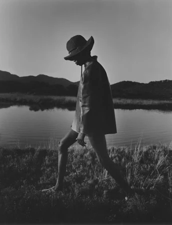 Model wearing a wide-brimmed hat and dark jacket walking by a calm lake with distant hills at dusk.