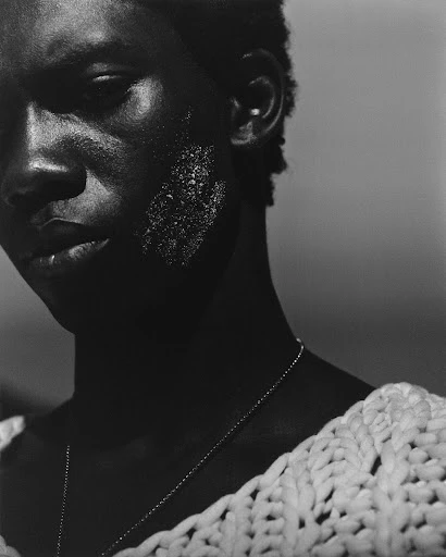 Close-up portrait of a Black male model with textured skin, wearing a knit top and necklace against a soft grey gradient backdrop.
