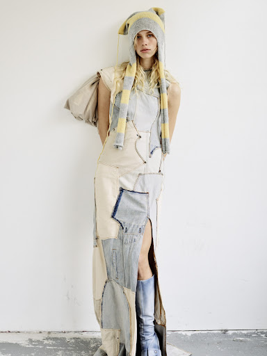 Woman in patchwork dress and beanie poses against a white wall in a studio. The dress has a thigh-high slit and blue boots complete the outfit.