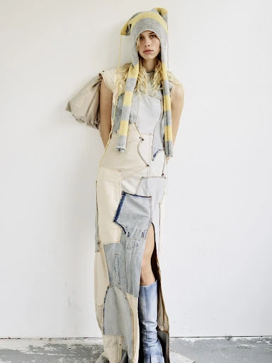 Woman in patchwork dress and beanie poses against a white wall in a studio. The dress has a thigh-high slit and blue boots complete the outfit.