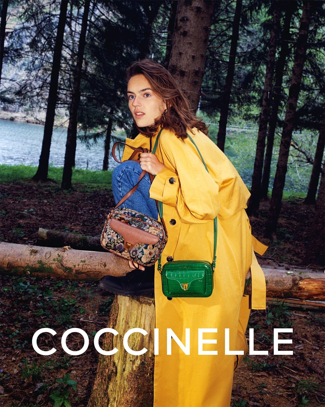 A fashionable adult female wearing a yellow coat and jeans poses in a wooded area carrying a couple of Coccinelle brand purses.