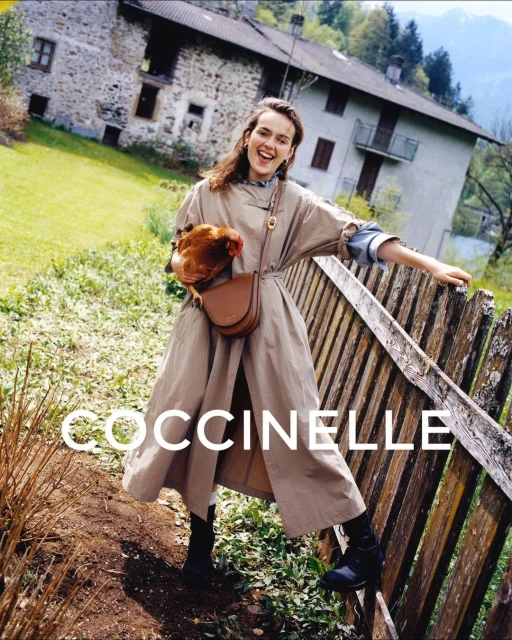 A smiling woman in a tan trench coat stands next to a wooden fence holding a tan leather purse and a brown chicken in a rustic outdoor setting.