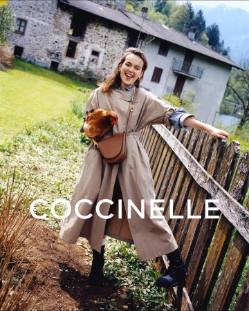 A smiling woman in a tan trench coat stands next to a wooden fence holding a tan leather purse and a brown chicken in a rustic outdoor setting.