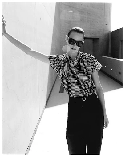 A stylish young woman with sunglasses leans against a wall outdoors. She wears a checkered top and dark pants with a belt in the sunlight.