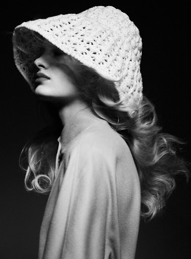 Black and white portrait of an adult woman with long wavy hair wearing a crochet hat in a studio setting with dark background.