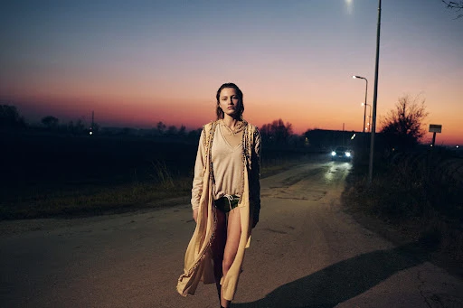 A woman in a beige robe stands on a road at twilight, with a car in the distance and a colorful sky at dusk or dawn.
