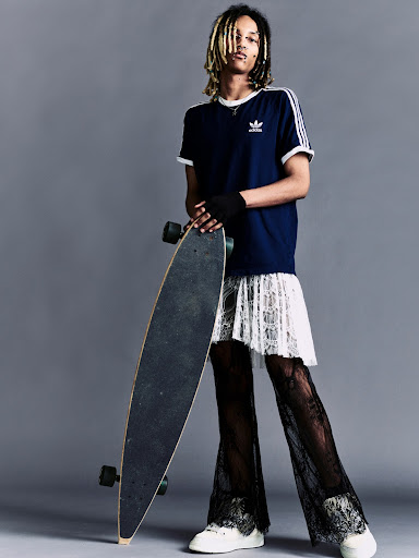 Full-shot of a person with dreadlocks wearing a navy t-shirt, lace skirt, and pants, holding a skateboard against a grey background.