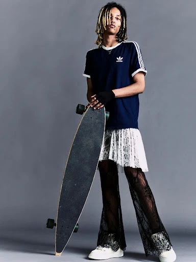 Full-shot of a person with dreadlocks wearing a navy t-shirt, lace skirt, and pants, holding a skateboard against a grey background.