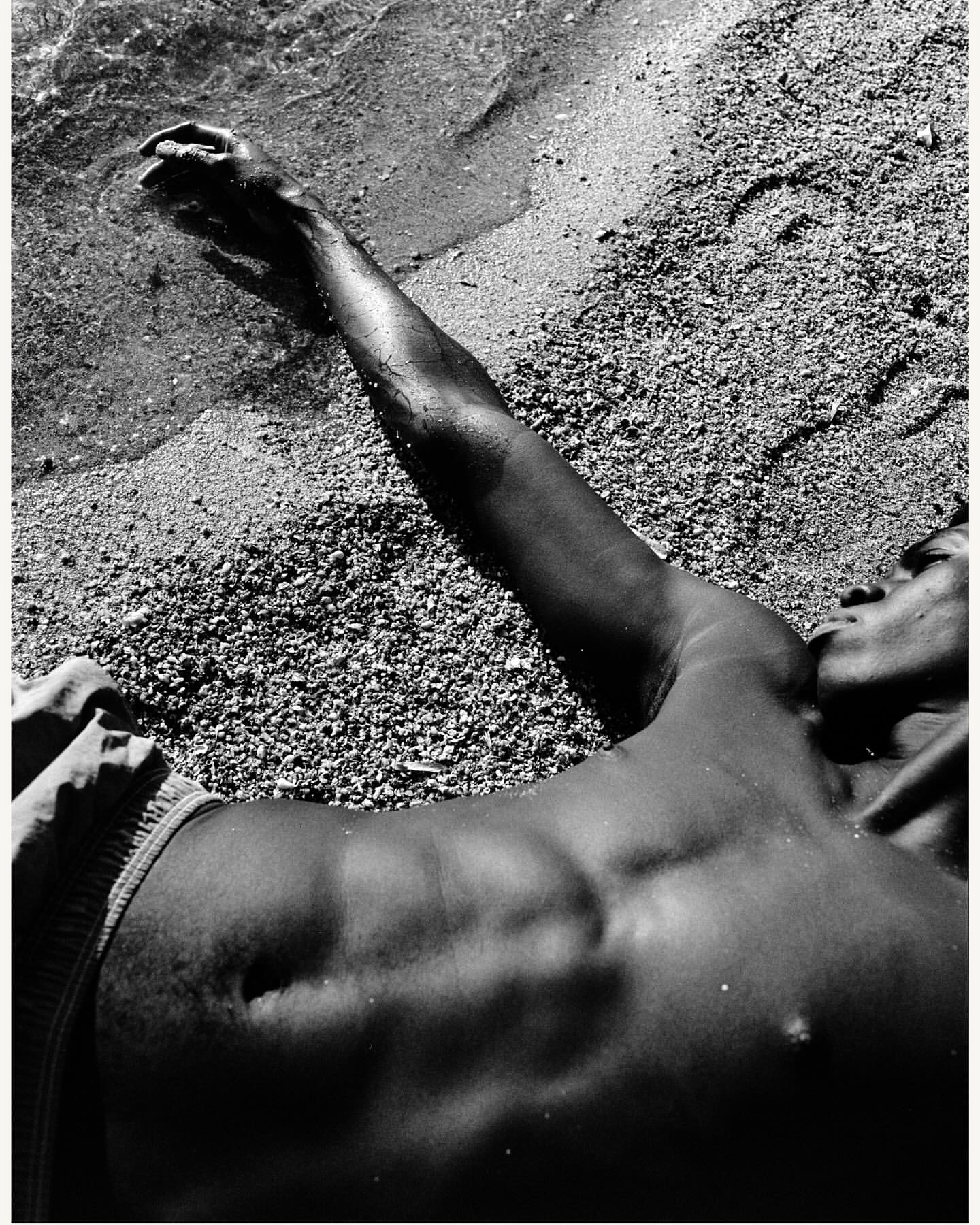 Monochrome photograph of an adult lying on a sandy beach with arm outstretched towards the ocean water lapping on the shore in daylight.
