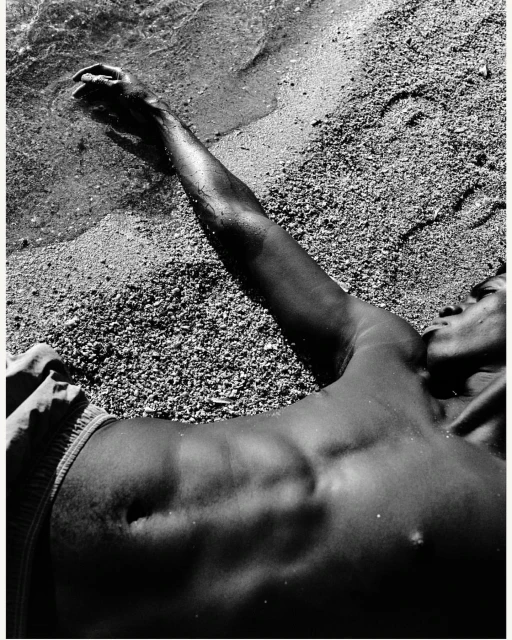 Monochrome photograph of an adult lying on a sandy beach with arm outstretched towards the ocean water lapping on the shore in daylight.